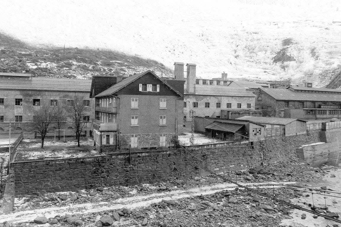 Photographie en noir et blanc de l'usine de Prémont vue depuis la rive de l'Arc : bâtiments de production, immeuble de logements et mur de soutènement, un câble traversant le premier plan