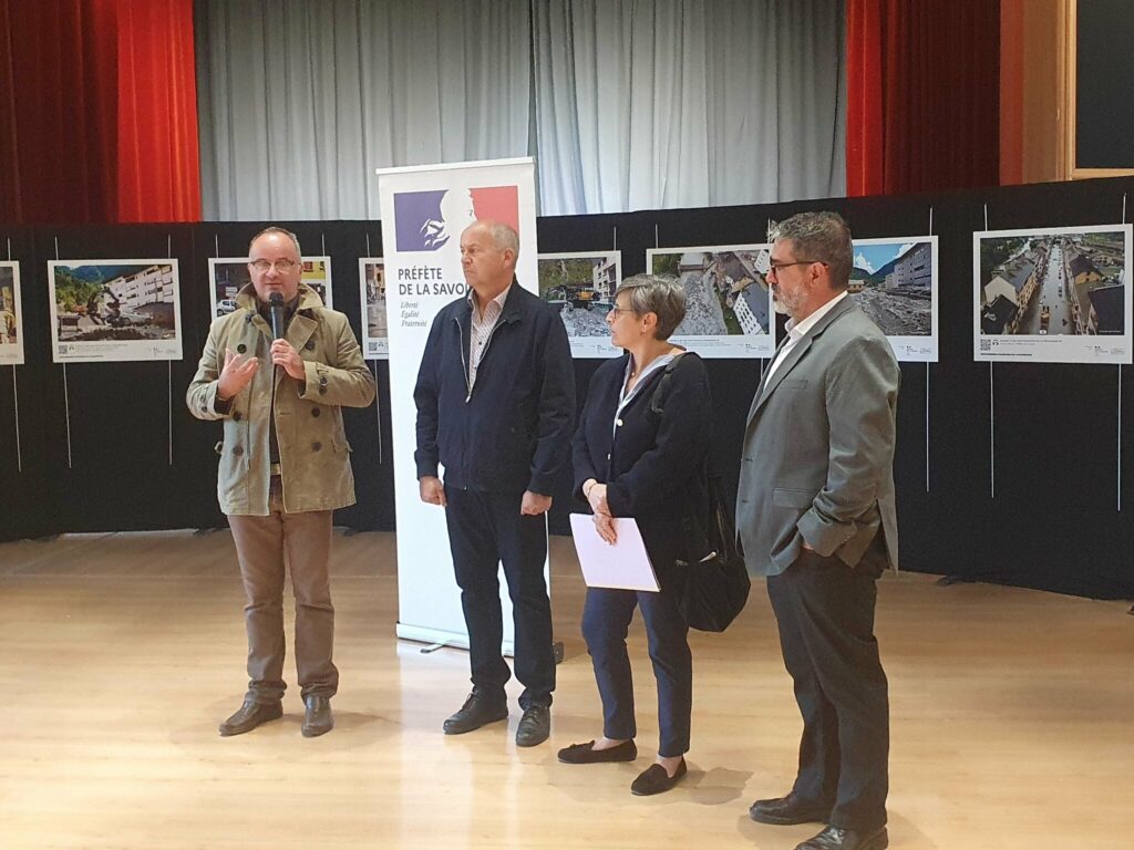Vernissage à la salle des fêtes de Fourneaux : François CHEMIN, maire de Fourneaux, parle au micro à côté d'Yves DURBET, de Magali SPANIOL et d'Humberto FERNANDES, devant le kakemono de la préfète de la Savoie et les photos de la crue exposées