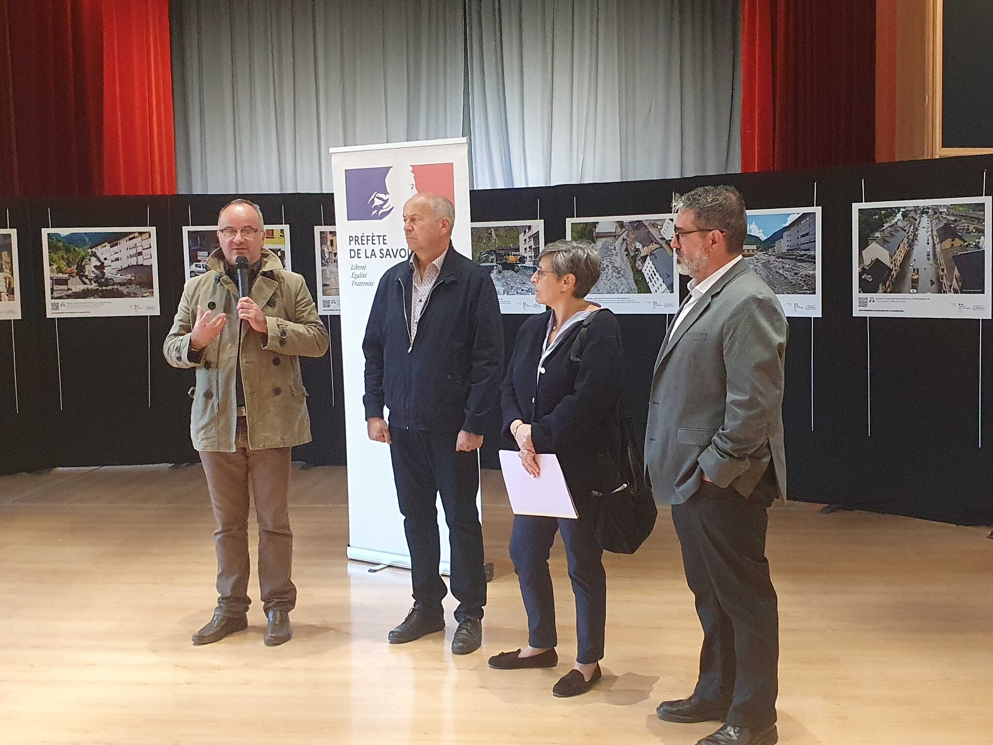 Vernissage à la salle des fêtes de Fourneaux : François CHEMIN, maire de Fourneaux, parle au micro à côté d'Yves DURBET, de Magali SPANIOL et d'Humberto FERNANDES, devant le kakemono de la préfète de la Savoie et les photos de la crue exposées